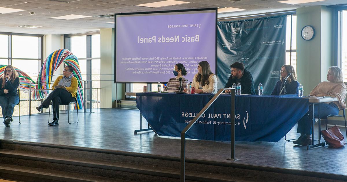 Several speakers at a table in front of a projection that reads basic needs panel